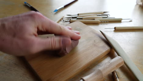 an artist student sculpting with soft brown modeling clay, squeezing and shaping it with his hands and fingers in an art studio with tools on the table