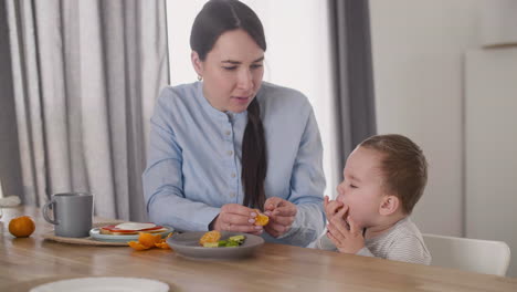 Mother-Feeding-Her-Little-Baby-Boy-With-Segments-Of-Clementine-While-Sitting-Together-At-Table-In-Living-Room