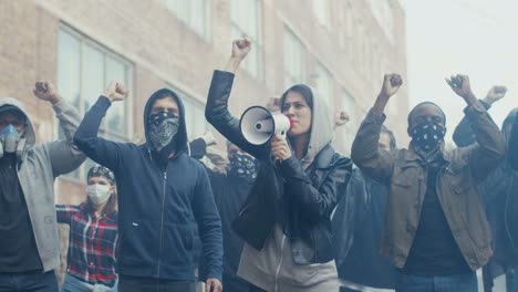 caucasian young woman in hood screaming mottos in megaphone. multiethnic male and female crowd of protesters shouting slogans and protesting. girl leading at manifestation for human rights.