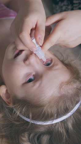 careful mommy drops little blonde girl eyes with new cure on soft beige bed in light room close upper view