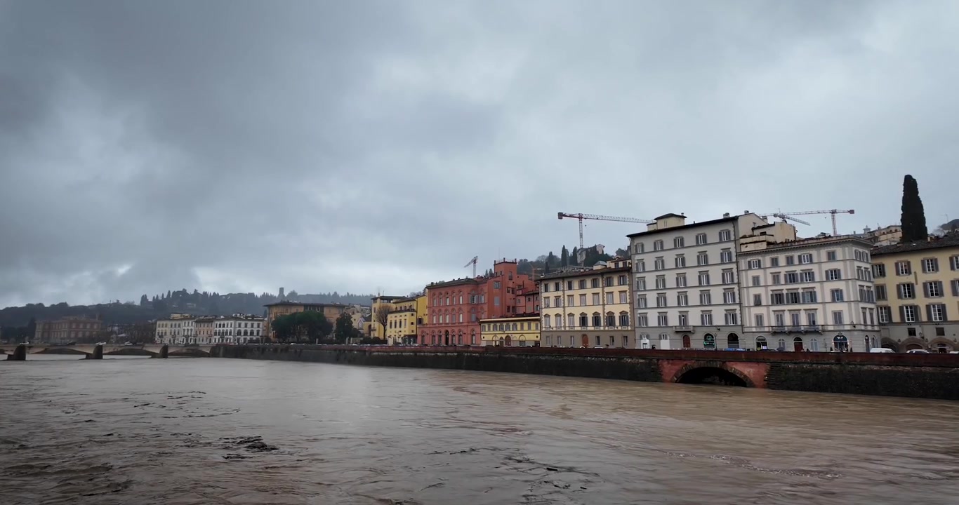 Vista panoramica del fiume Arno a Firenze durante una giornata tempestosa, con acque alluvionali fangose, edifici storici lungo il fiume e nuvole drammatiche sulla città toscana.