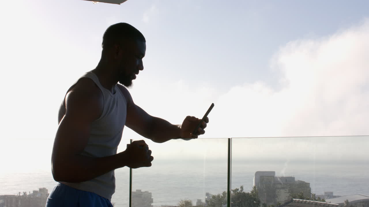 Strong and fit African American man using smartphone while exercising on balcony at home