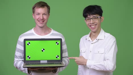 2人の若い多民族のビジネスマンが指を指さしながらカメラにラップトップを示しています