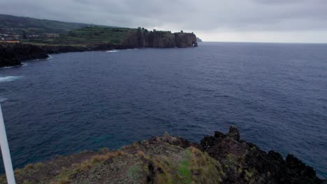 Flag-of-capelas-village,-drone-flying-towards-the-seashore-of-São-Miguel-Island,-Portugal