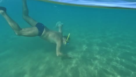 split underwater view of fisherman diving with mask and snorkel looking for razor clams hidden in seabed while holding bag full of sea food in crystalline sea water