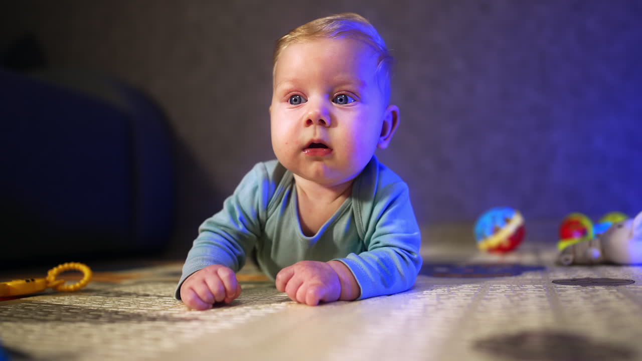 Little baby boy with fair hair and big eyes looks with interest around. Infant kid lies on the floor at home.