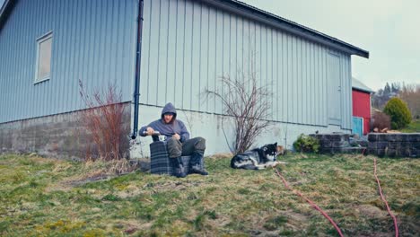a man sits outside a house, pouring a drink into a mug while a dog rests nearby on the grass - static shot