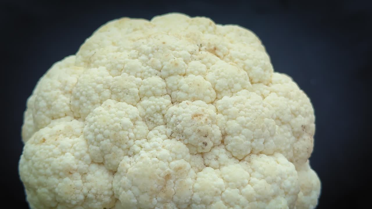 Primo piano su un giradischi con una testa di cavolfiore fresca che ruotaFluidamente su un piatto; i suoi fiori dettagliati catturano la luce soffusa mentre il movimento delicato ne rivela la consistenza e la forma naturale.