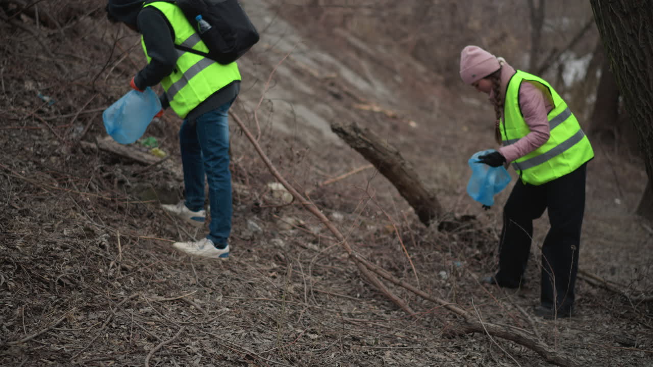 Two volunteers in safety vests collecting litter in wooded riverside area during environmental cleanup, carrying blue plastic bags and wearing warm clothing while working together to remove waste