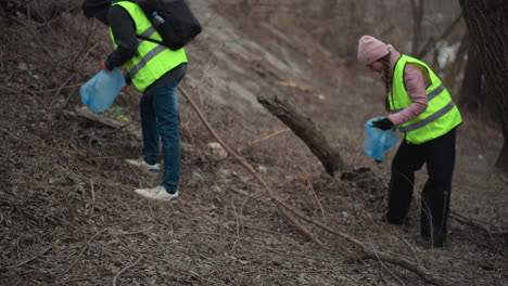 two volunteers in safety vests collecting litter in wooded riverside area during environmental cleanup, carrying blue plastic bags and wearing warm clothing while working together to remove waste