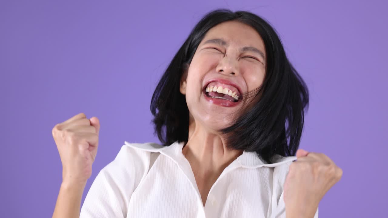 Woman in white shirt celebrates energetically, smiling and raising fists, against a purple background