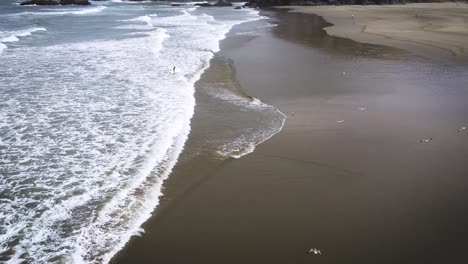 Surfista-Saliendo-Del-Océano-Mientras-Un-Dron-Revela-Las-Majestuosas-Escenas-De-La-Playa-De-Totoritas,-Perú