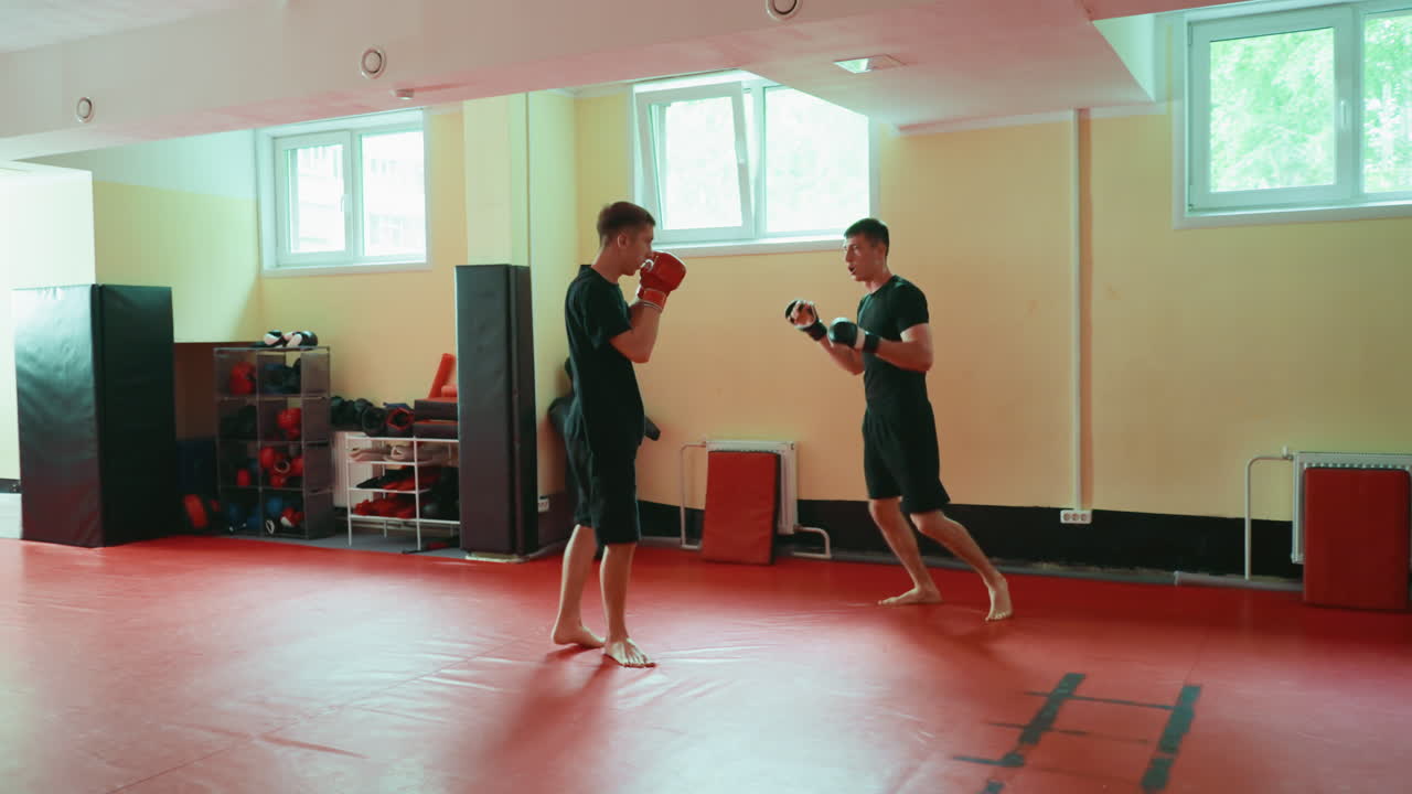Two fighters sparring in gym on red mat, barefoot, wearing gloves, facing each other in combat stance, practicing martial arts techniques, focusing on agility, strength, discipline under natural window light