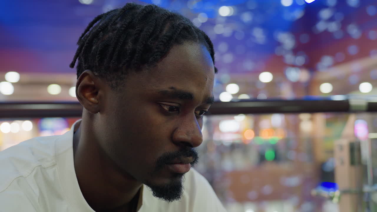 Close up of man seated in shopping mall wearing white t-shirt looking contemplative and thoughtful with focused expression as colorful lights blur in background