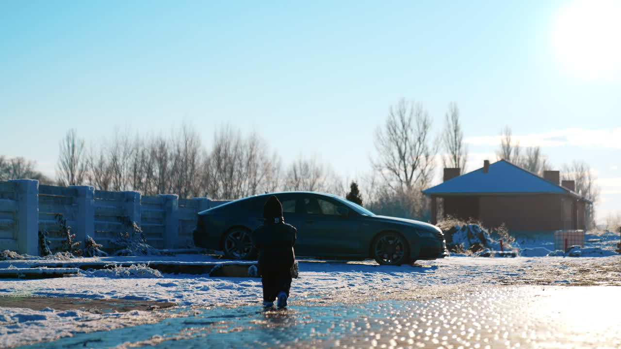 Rear view of the little toddler boy wearing black clothes and cap. Baby boy walks outdoors in winter in the yard.