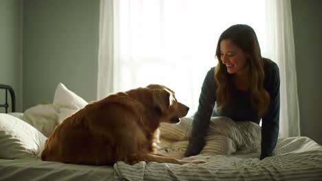 a woman enjoys a heartwarming interaction with her golden retriever while seated on a bed in a softly lit bedroom. the warmth of the morning sun enhances their joyful connection.