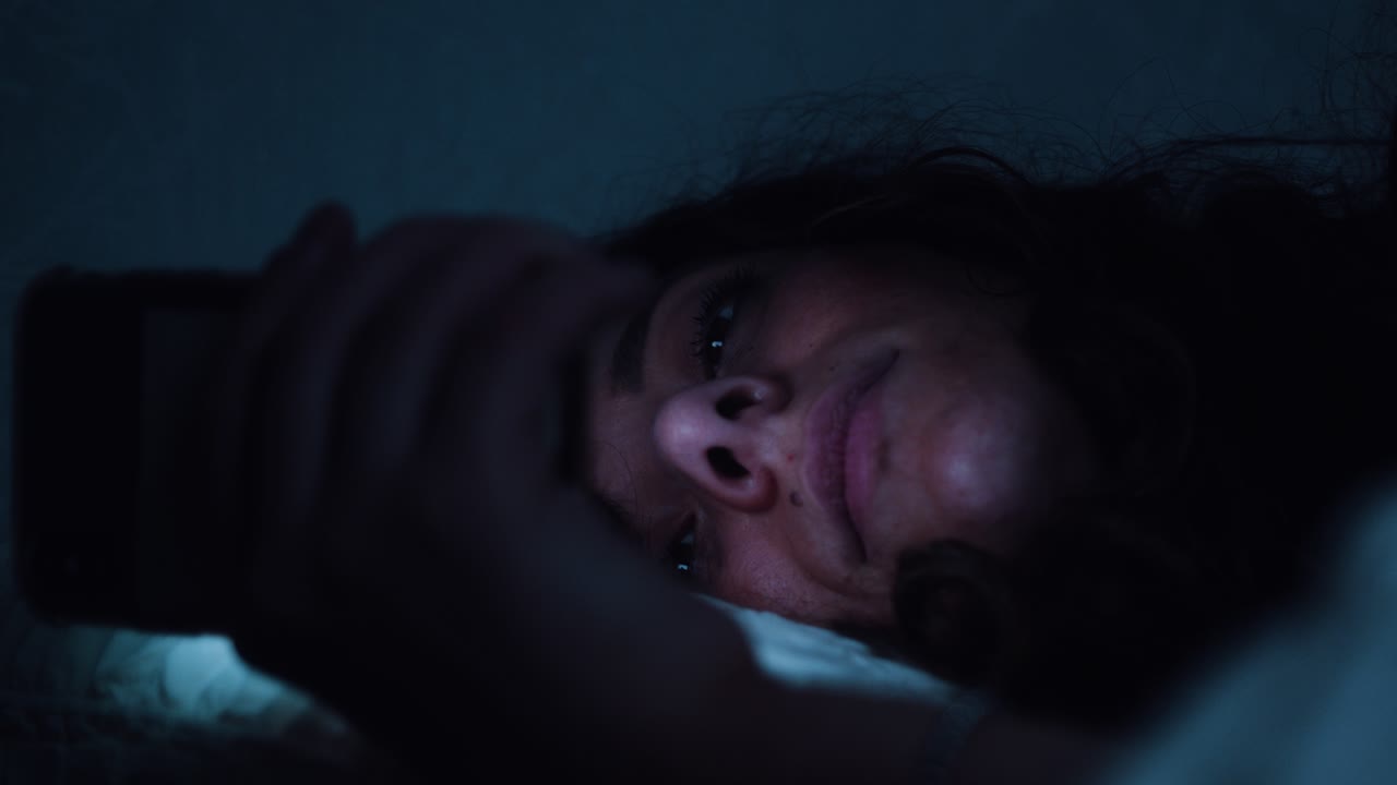 una mujer cansada lucha por mantener los ojos abiertos mientras usa un teléfono inteligente en la cama.