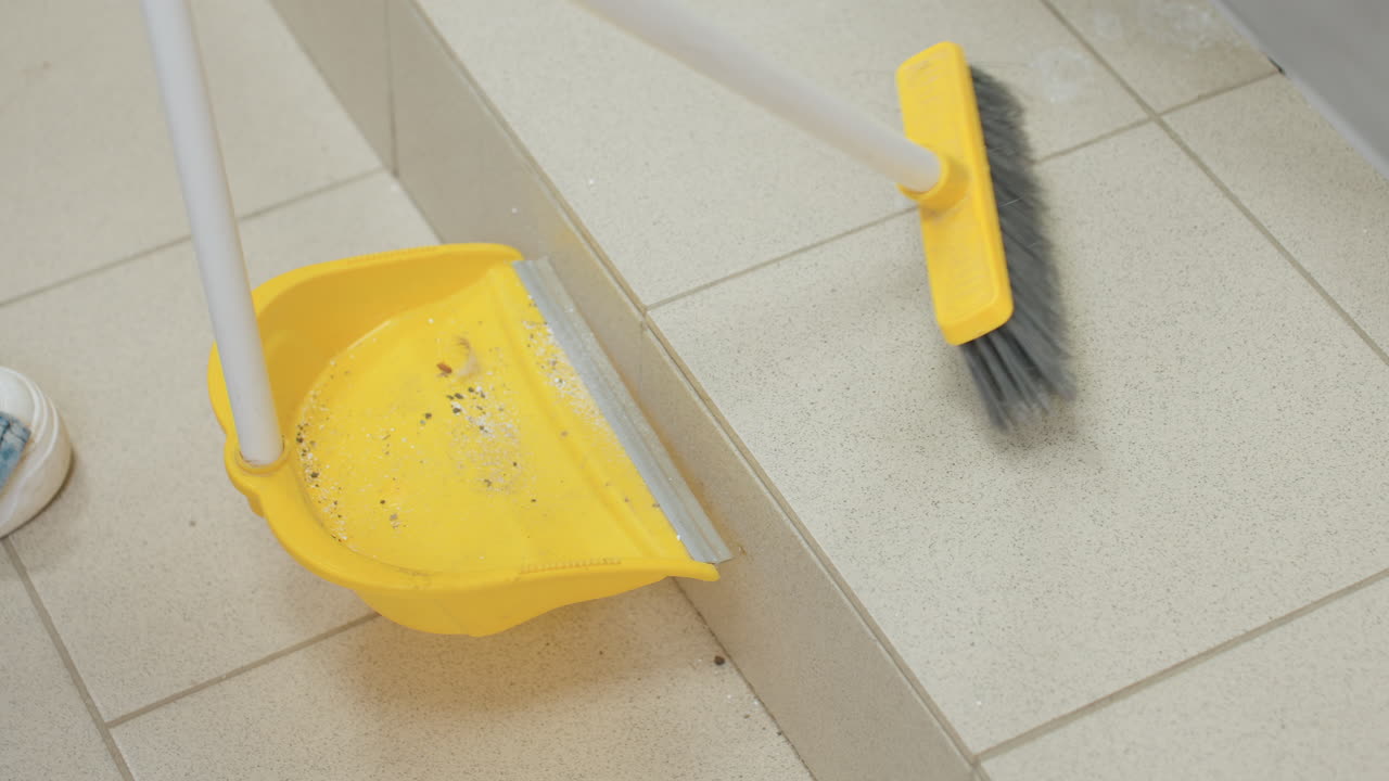 High angle view shows female in white sneaker sweeping dirt toward pan with yellow broom on tiled floor beside stainless washer,bristles push debris during maintenance inside bright laundry room