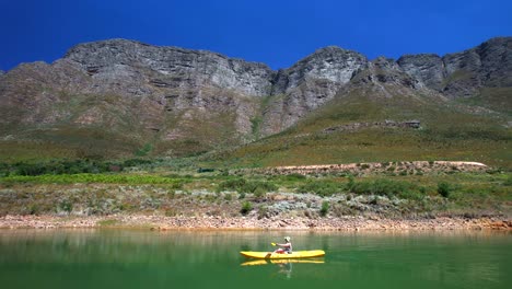 person paddling a bright yellow canoe from right to left across turquoise water. majestic mountains rise in the background, mirrored perfectly in the still pond