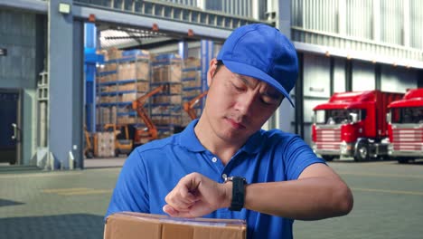 close up of asian male courier in blue uniform checking time on his watch while holding a carton, outside of logistics distributions warehouse