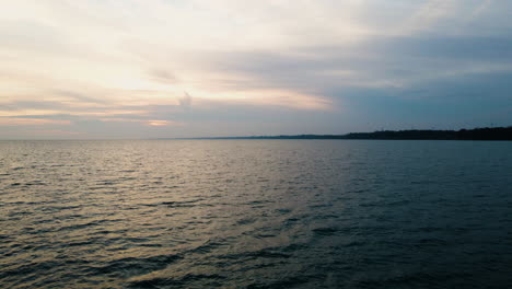 Golden-hour-light-diffused-by-clouds-reflects-on-calm-water-ripples,-silhouette-of-land-in-distance-at-sunset