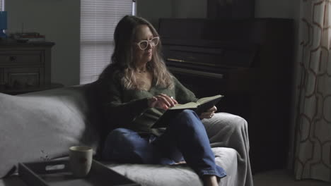 beautiful adult woman wearing eyeglasses reading book sitting in the couch at home