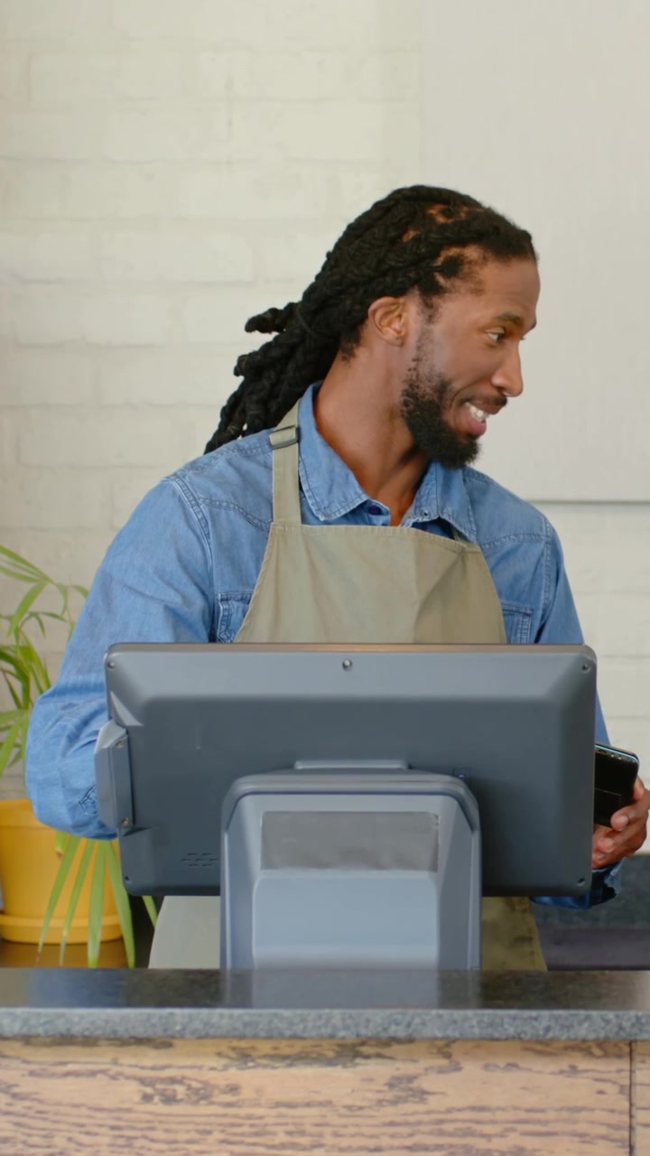 Vertical video: Customer handing device, cashier in beige apron tapping POS at counter, charging