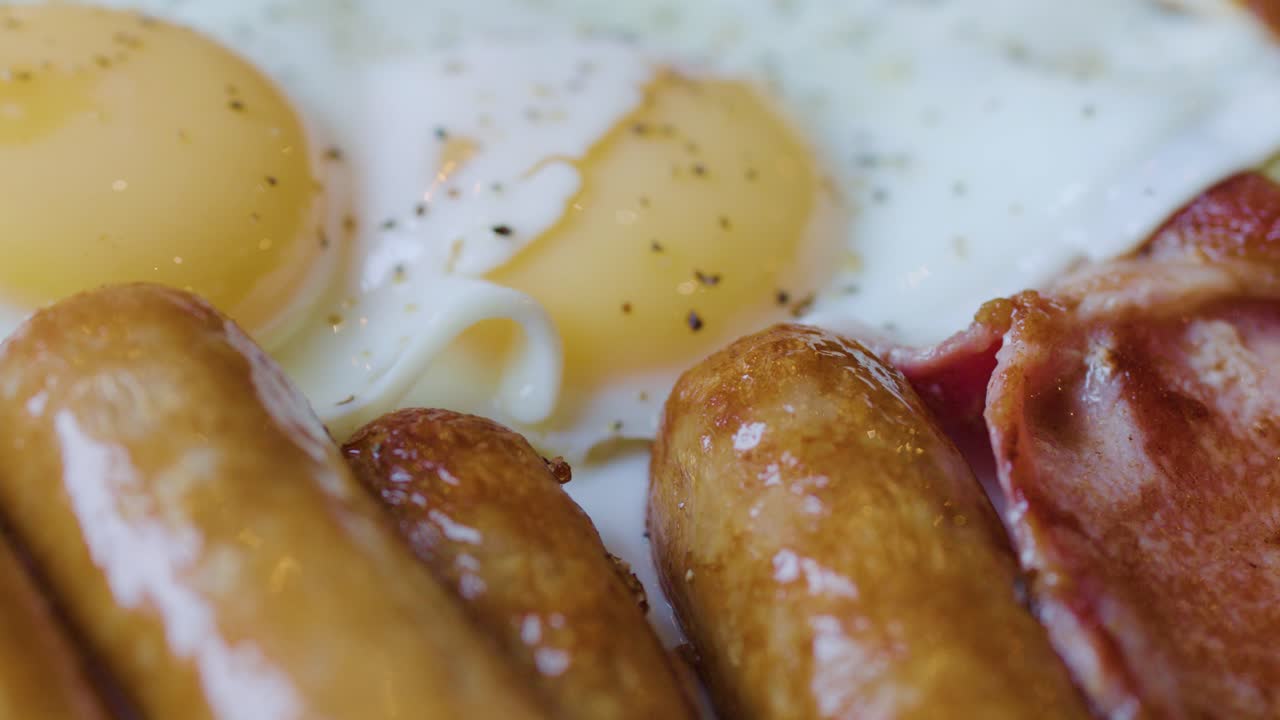 Hand cuts sausage with knife and fork beside eggs and bacon, natural daylight, macro shot