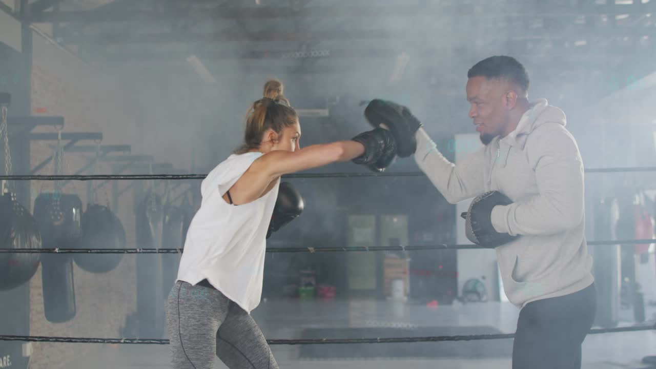 Female gloved trainee throwing punch combinations while trainer cueing with mitts in boxing ring