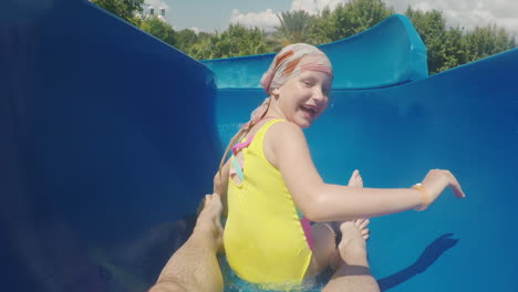 una familia feliz con un niño está montando un tobogán de agua riendo agitando sus manos a la cámara lenta m