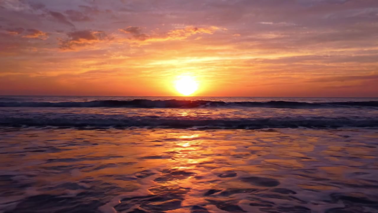 En smuk solnedgang over rolige havbølger, der reflekterer en rig palet af orange, lilla og lyserøde nuancer, hvilket skaber en fredfyldt og rolig strandatmosfære i skumringen