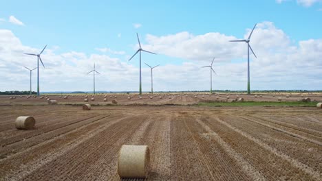 Luftaufnahmen-Zeigen-Die-Ruhige-Schönheit-Der-In-Bewegung-Befindlichen-Windturbinen-Auf-Dem-Kürzlich-Abgeernteten-Feld-Eines-Landwirts-Aus-Lincolnshire,-Wo-Goldene-Heuballen-Den-Vordergrund-Schmücken