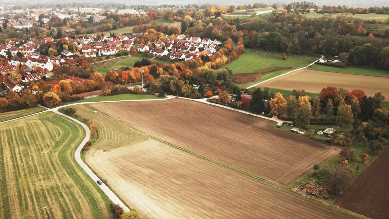 Camera glides forward over dense golden treetops and patchwork fields, approaching a picturesque German village nestled in seasonal colors