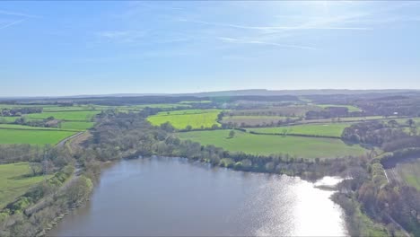 Drone ascends over large pond in Elsecar countryside with fields stretching beyond, UK