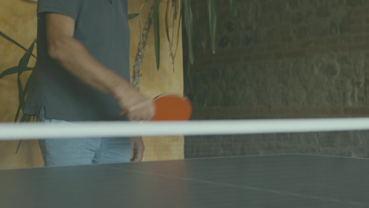 A man hits the white ball in ping pong during a game, detail of the net and racket in slow motion, the table is in a tavern and plants and a yellow brick wall can be seen in the background