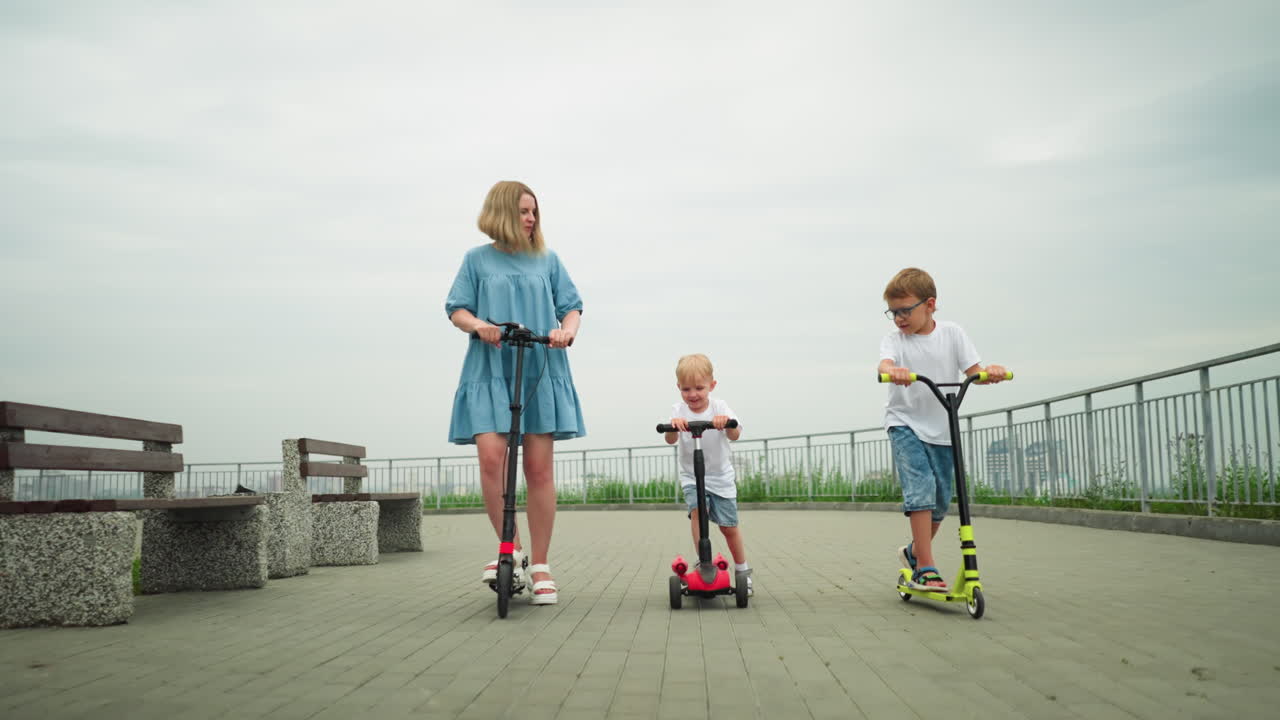 A mother and her two children ride scooters together in an open park area, with an iron rail by the side and benches around