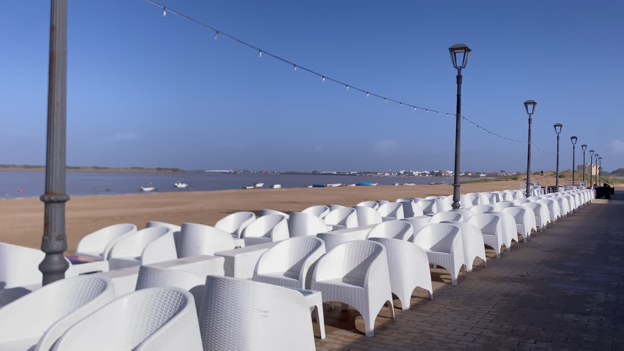 White chairs and tables line a coastal promenade, presenting an inviting setting for outdoor dining by the beach