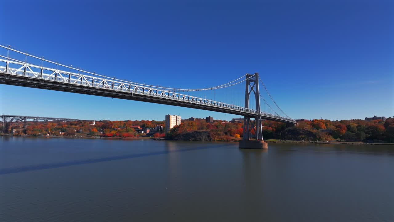 An aerial view over the Hudson River showing the Mid-Hudson Bridge on a sunny day in autumn. The camera dolly in over the calm water towards the bridge with Poughkeepsie, New York in the background.