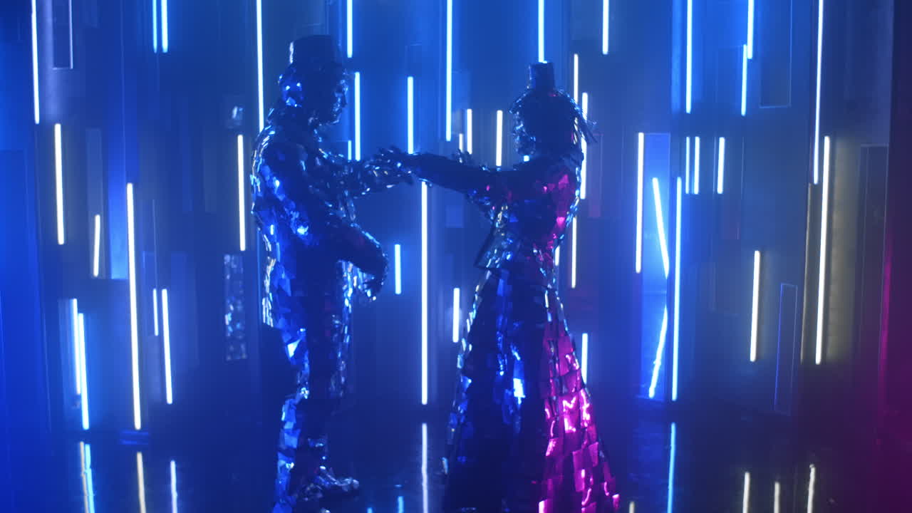 Two robot dancers in glittering costumes dance against a neon wall drawing closer to each other.