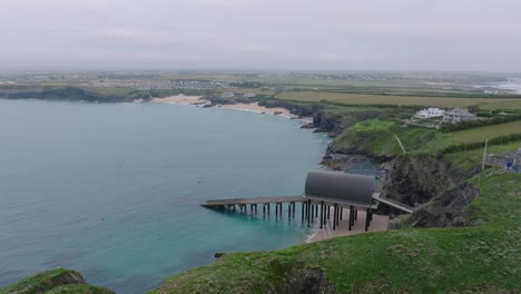 craggy sea cliffs with waves breaking. camera flight over revealing rnli lifeboat station with slipway next to clean sandy beach. mother ivey's bay, cornwall, uk.