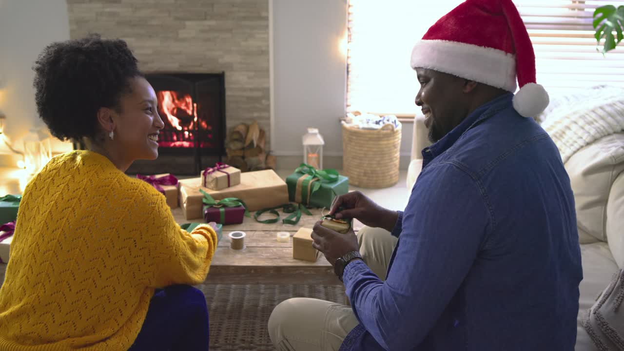 With man entering wearing Santa hat, diverse couple wrapping holiday gifts on floor by fireplace