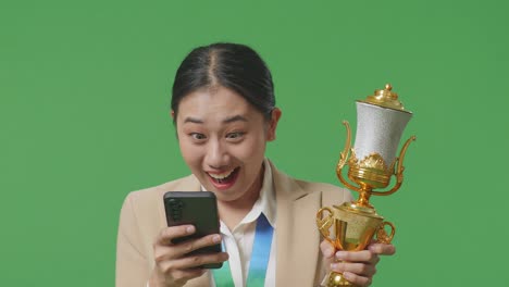 close up of asian business woman in a suit with a gold medal and trophy looking at smartphone then saying wow on green screen background in the studio