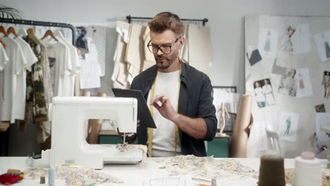 Man-Tailor-In-Glasses-Sitting-At-The-Table-With-A-Sewing-Machine-And-Using-Tablet,-Then-Smiles-At-Camera