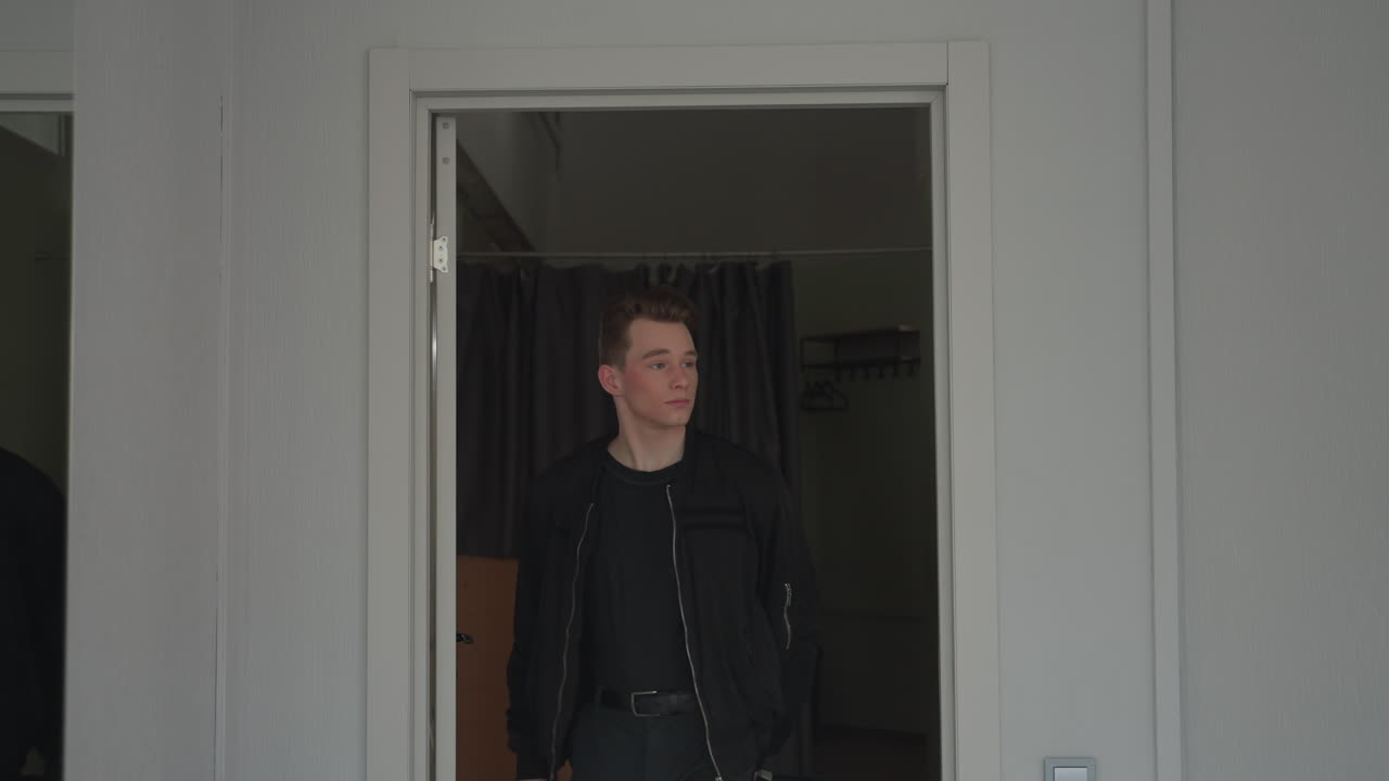 Student wearing black jacket exits modern apartment room through white paneled door stepping into bright hallway with pale gray walls light switch wooden floor curtain and clothes rack behind door