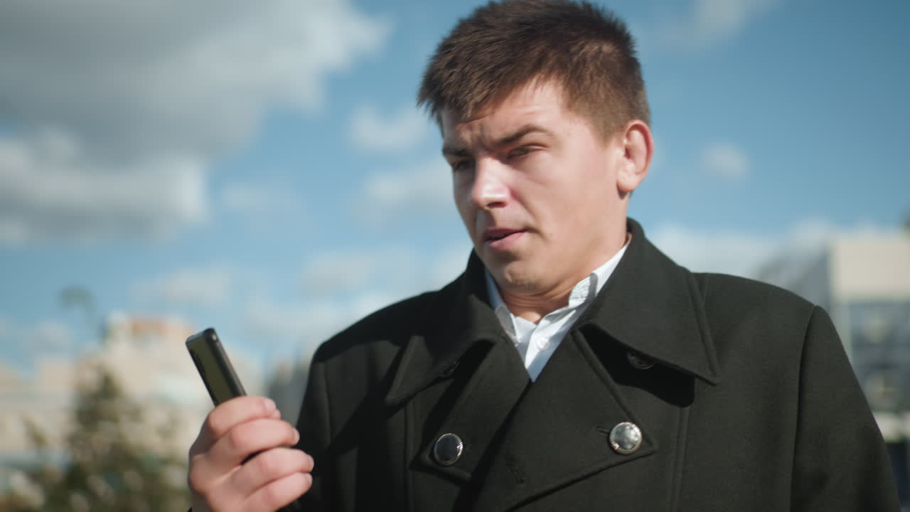 검은 코트를 입은 남자가 야외에서 전화를 하고, 대화에 반응하는 동안 불만으로 머리를 흔들고, 도시 배경에는 현대적인 건물, 초록색, 은 파란 하늘이 포함되어 있습니다.