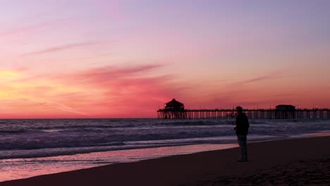 Un-Hombre-Disfruta-De-Sus-Vacaciones-En-La-Playa-Durante-Una-Hermosa-Puesta-De-Sol-Roja,-Púrpura,-Mandarina,-Rosa-Y-Azul-Con-El-Muelle-De-Huntington-Beach-Al-Fondo-En-Surf-City-USA-California