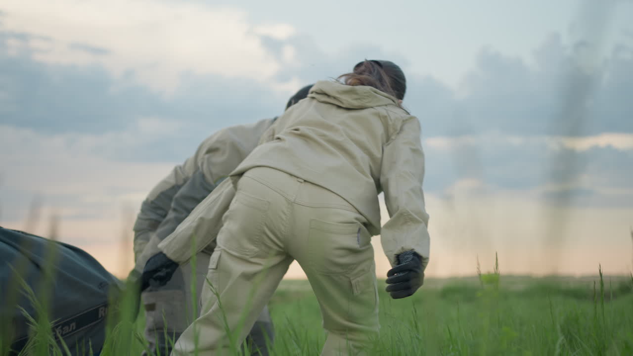 Man and woman wearing beige uniforms drag large blue balloon equipment through lush tall grass, working together in open field at dusk under cloudy sky, highlighting effort and rural teamwork