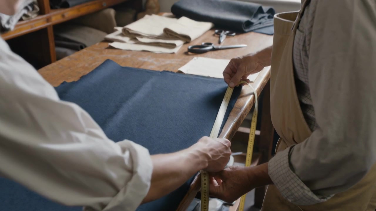Pulling tape senior tailor in apron guiding apprentice aligning tape on fabric at bench for cutting