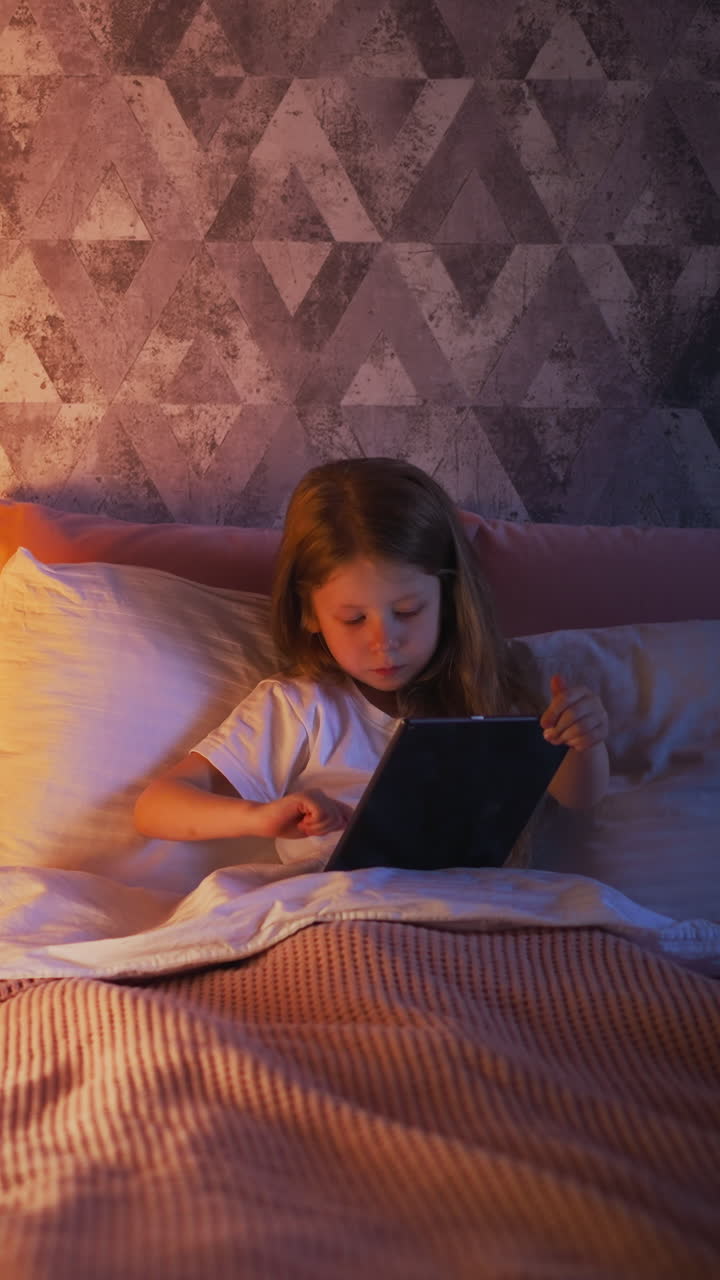 Long haired kid girl touches screen of tablet lying in bed on pillows under warm blanket by play tent with night lamp in semidark room slow motion