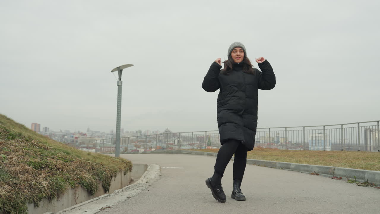 Joyful female moves by river. Energetic lady dancing on hilltop trail during sunset evening. Woman performing lively dance steps along riverside pathway with panoramic city and river views